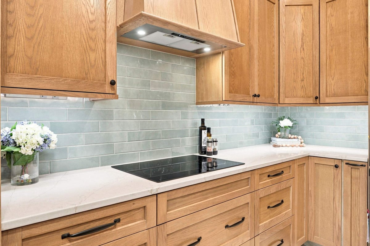 Wood range hood above an electric cooktop with blue-green subway tile backsplash and light countertops.