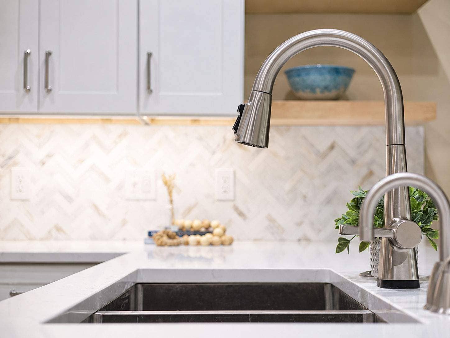 Brushed nickel kitchen faucet above a stainless sink with white cabinets and a softly lit tile backsplash.