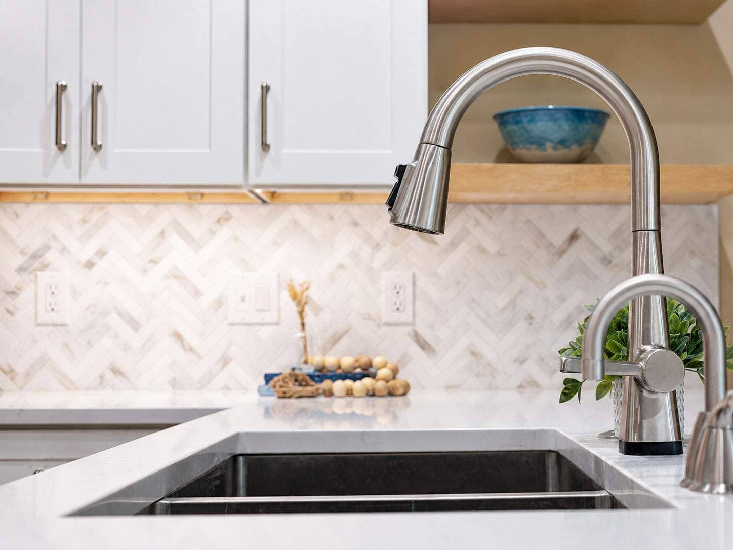 Brushed nickel pull-down faucet over a stainless sink with white cabinets and herringbone backsplash.