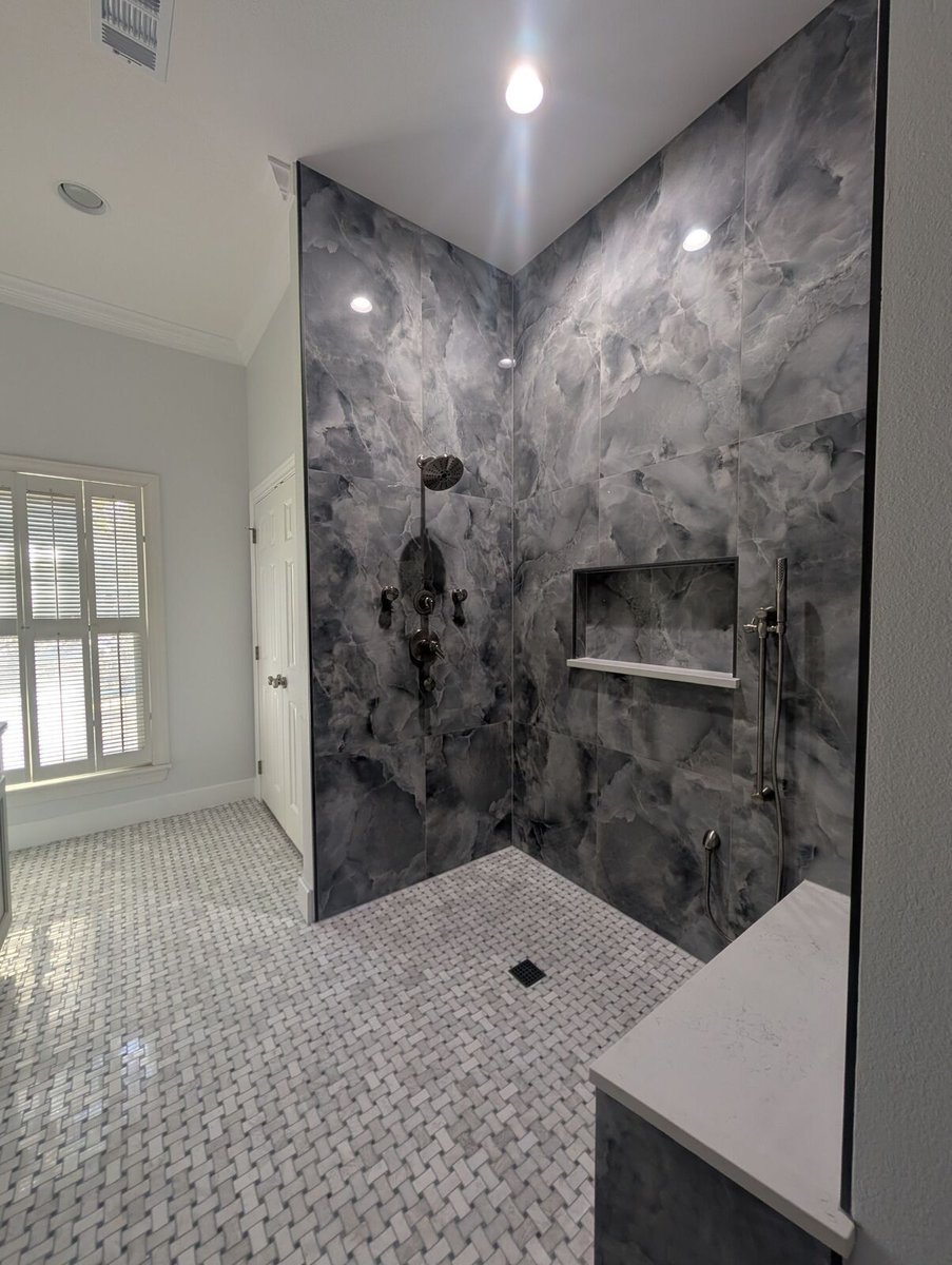 Finished bathroom shower with dark gray tile, glass panels, shower niche, bench and bright window light.