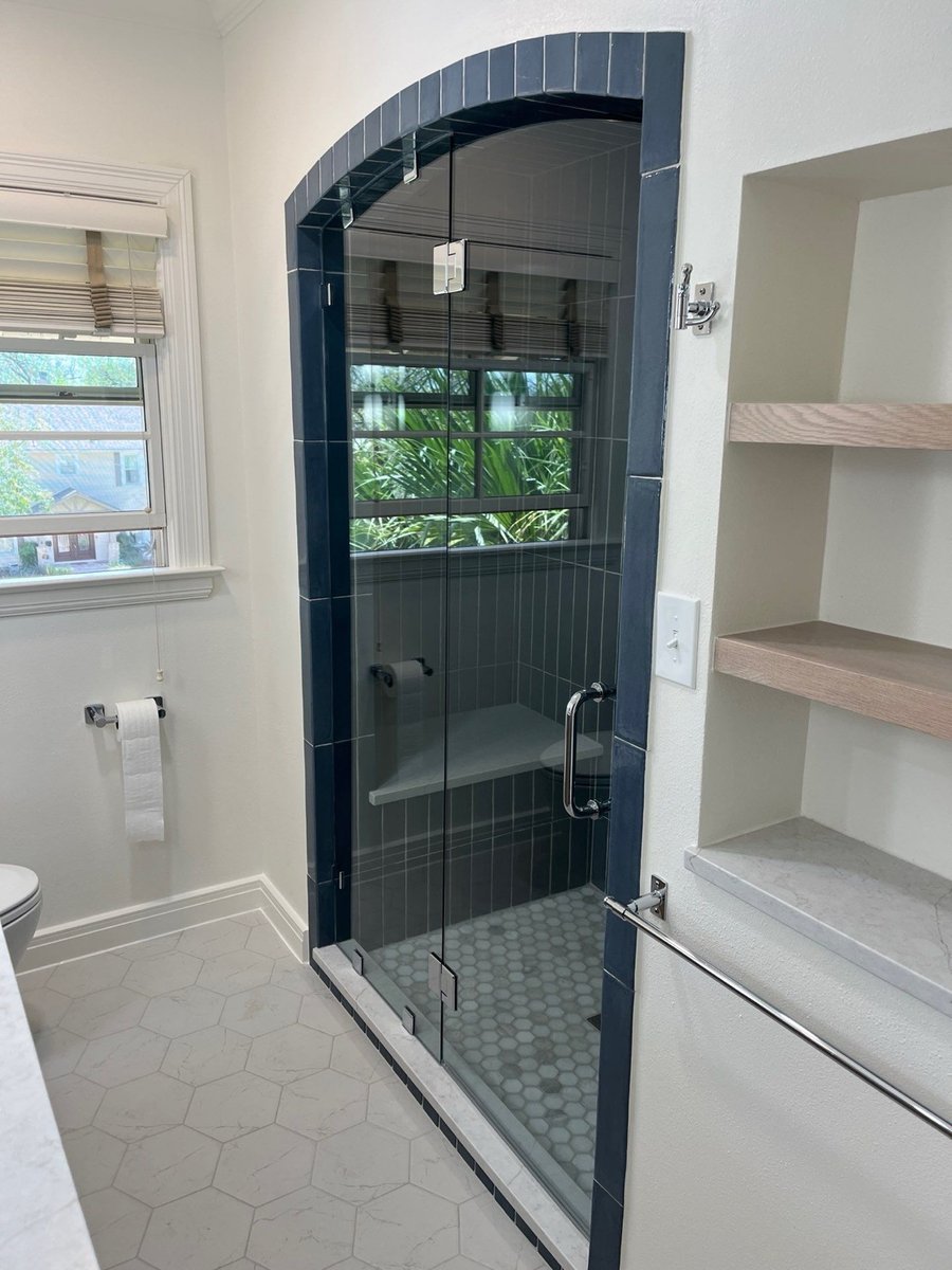 Arched glass shower with navy tile beside a window and white floating shelves