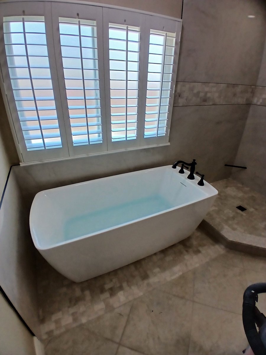 Freestanding white tub by shuttered window with marble tile and black faucet