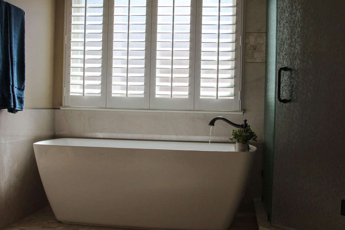 Freestanding white tub filling by a shuttered window beside glass shower