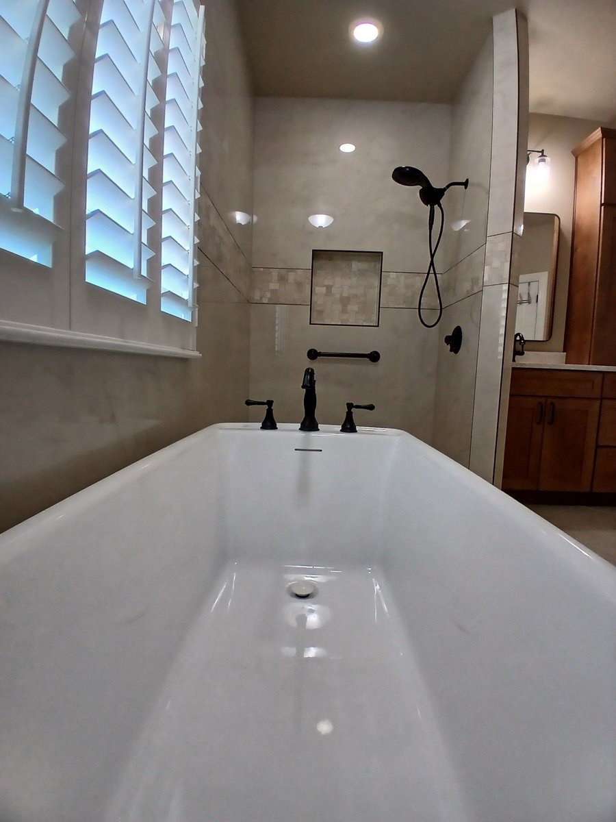 Freestanding tub with black faucet facing marble shower and maple vanity