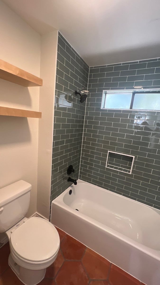 Remodeled tub shower with sage green subway tile, black fixtures, and terracotta hex floor