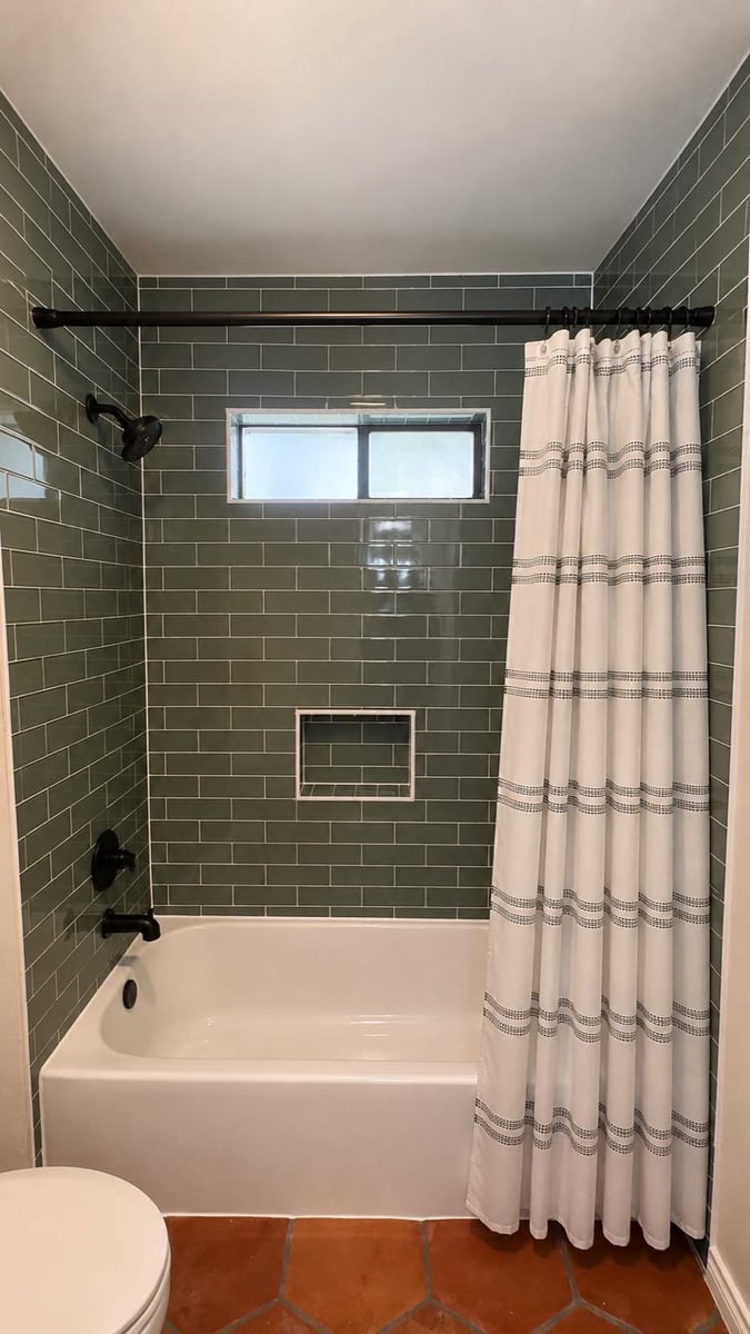 Green subway tile shower with white striped curtain on a black rod and terracotta floor