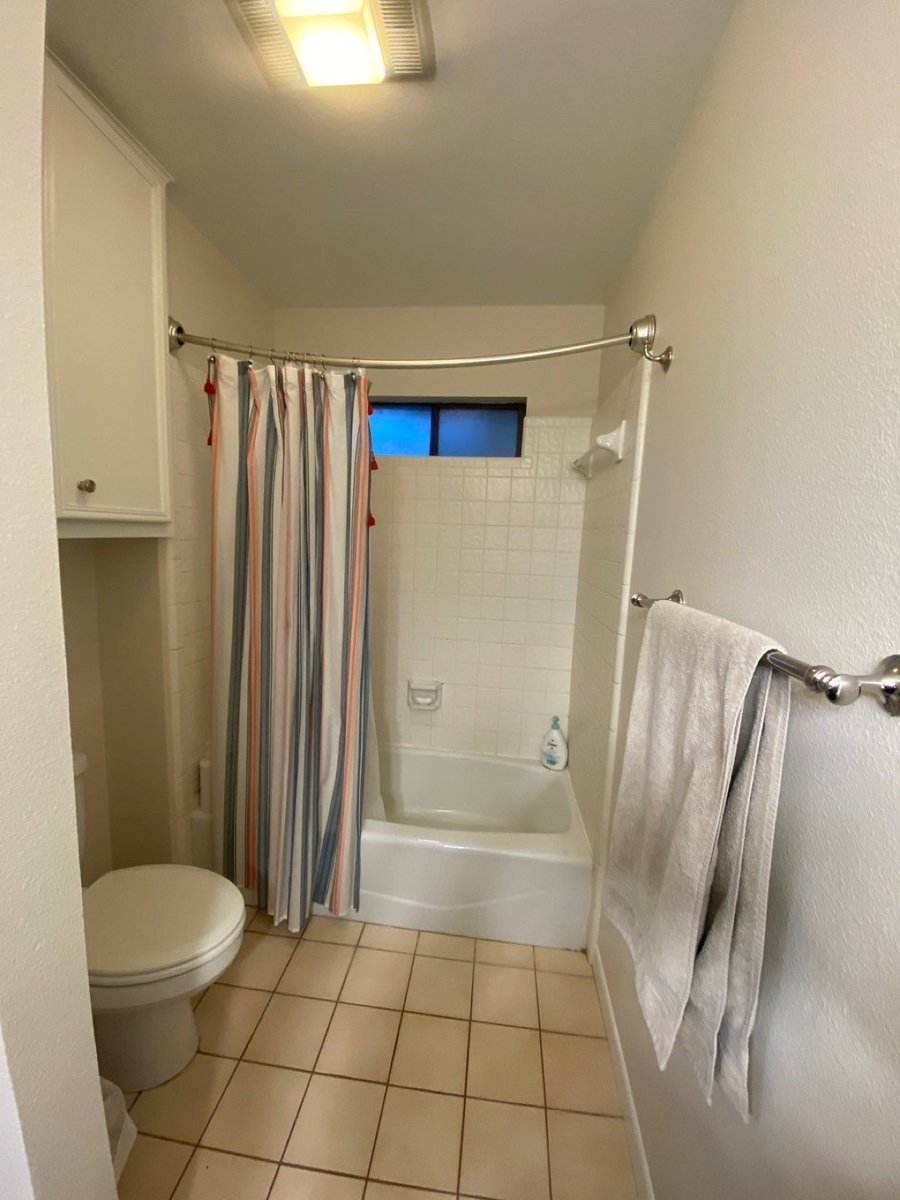 Original narrow bathroom with white tile tub surround, striped curtain, and beige floor tile before remodel