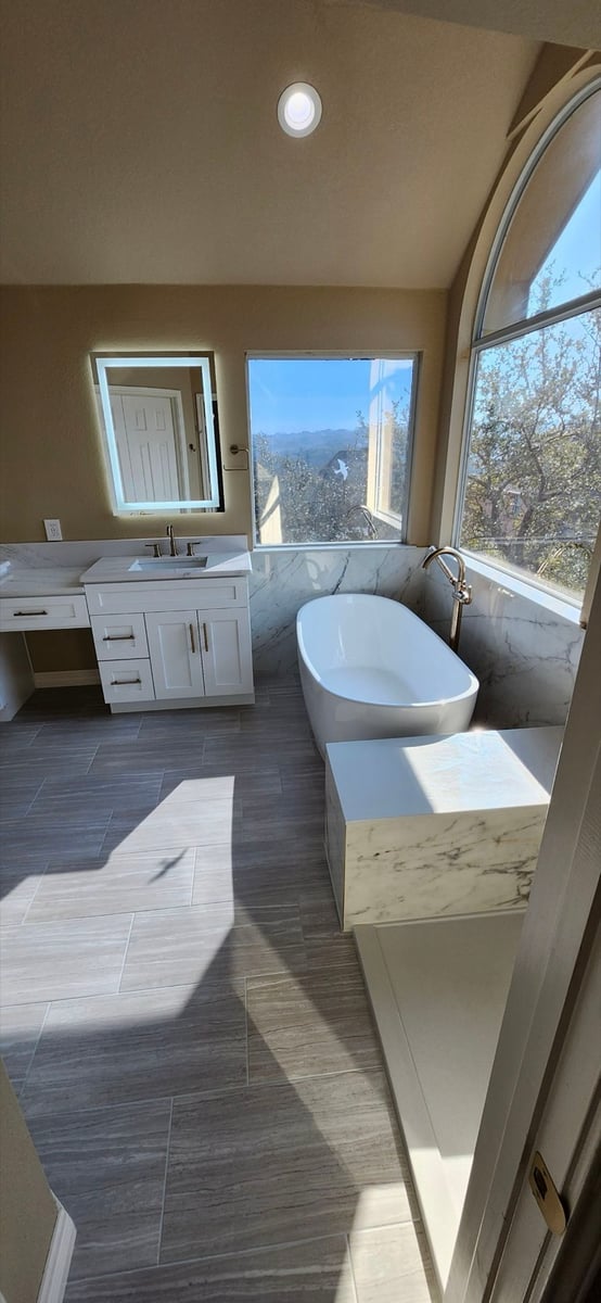 White vanity and freestanding tub by arched window with hill views