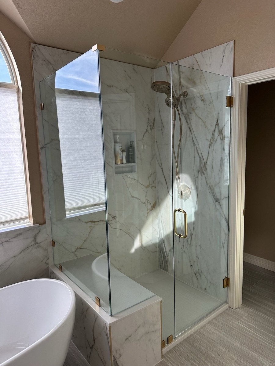 Glass-enclosed marble shower with bench, niche, and brass fixtures beside freestanding tub