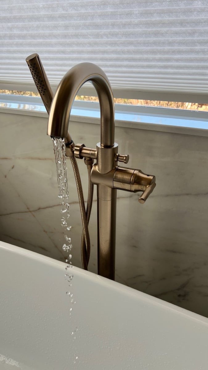 Aged brass floor-mounted tub filler running water beside a marble-walled tub