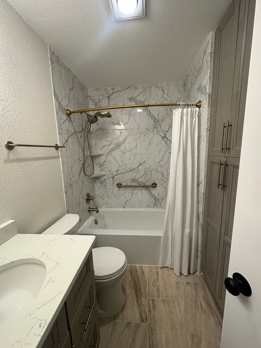 Guest bath with tub-shower, marble-look walls, gray vanity, and gold shower rod