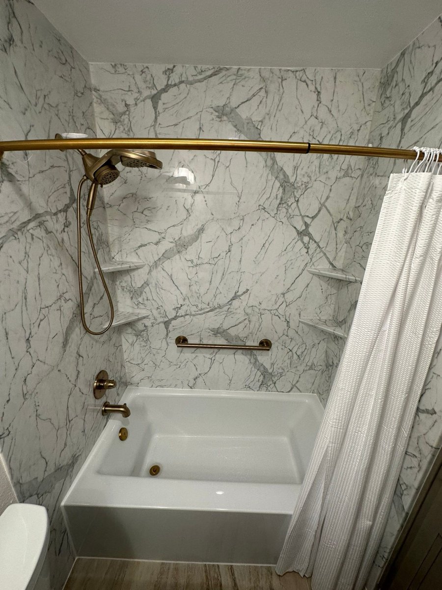 White tub with brushed gold fixtures, corner shelves, and curved shower rod on marble walls