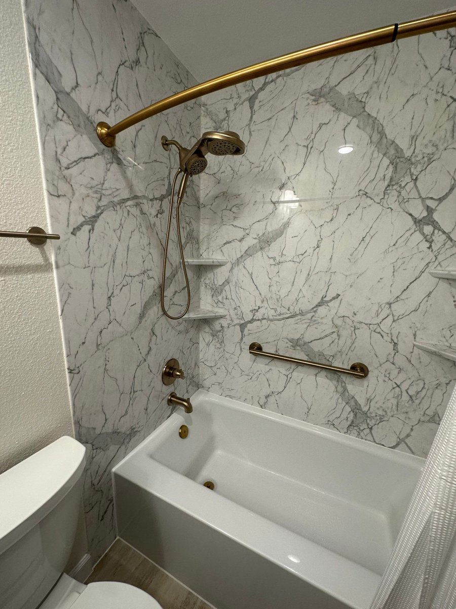 Tub-shower with gold handheld showerhead and grab bar on marble-look wall panels