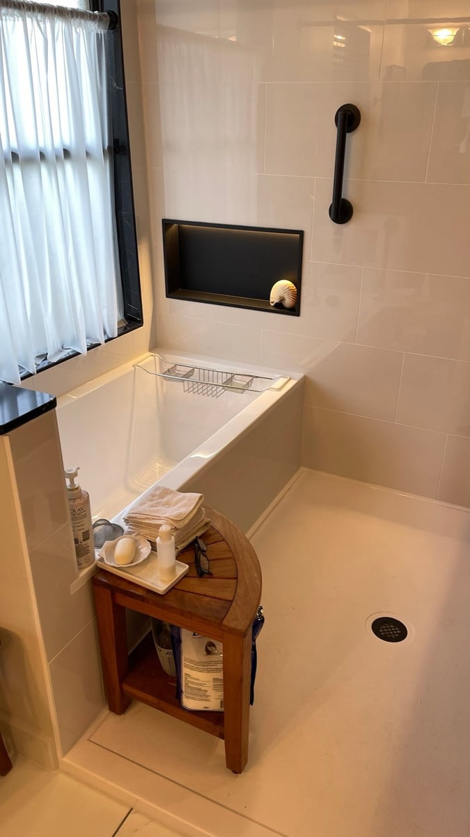 White tub beside black marble shower with lit niche and teak bath stool