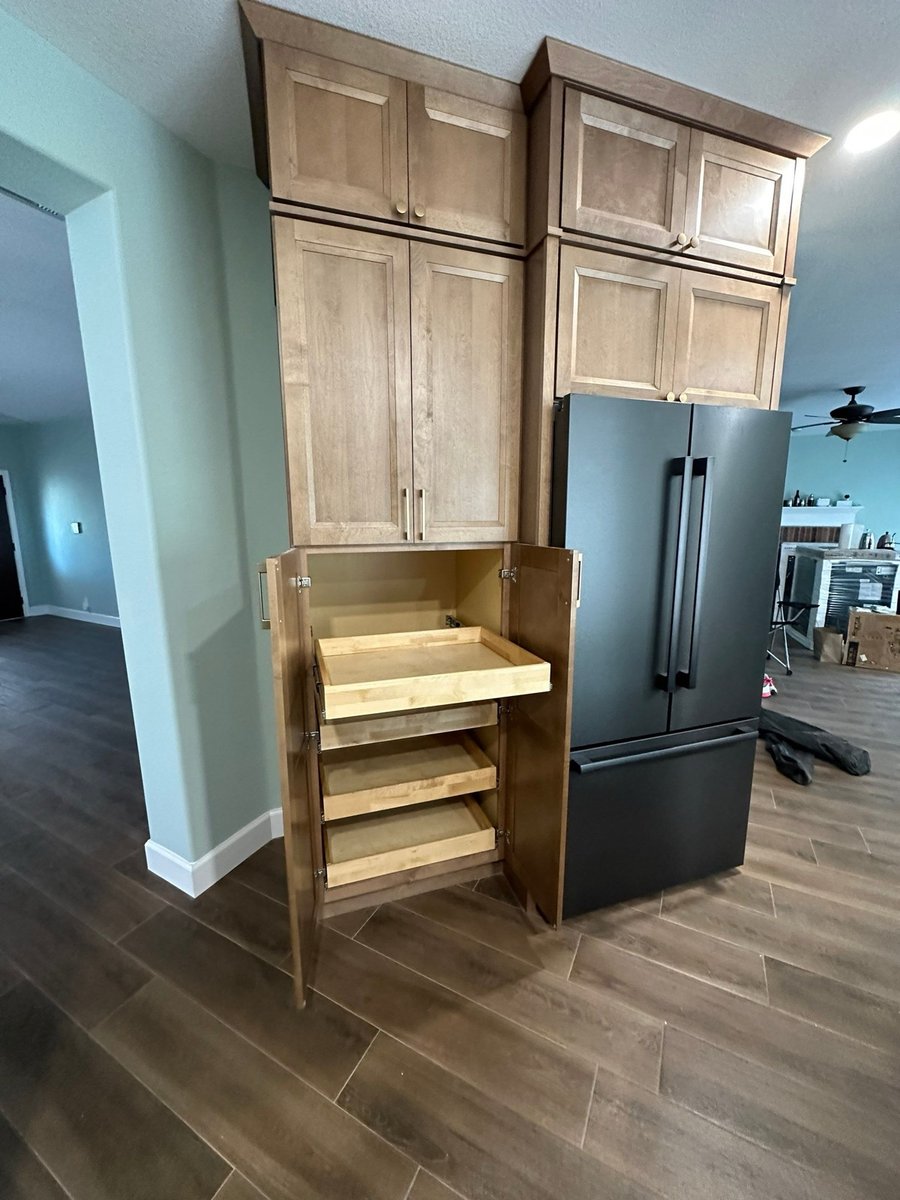 Open pantry cabinet revealing wood pull-out shelves next to black refrigerator