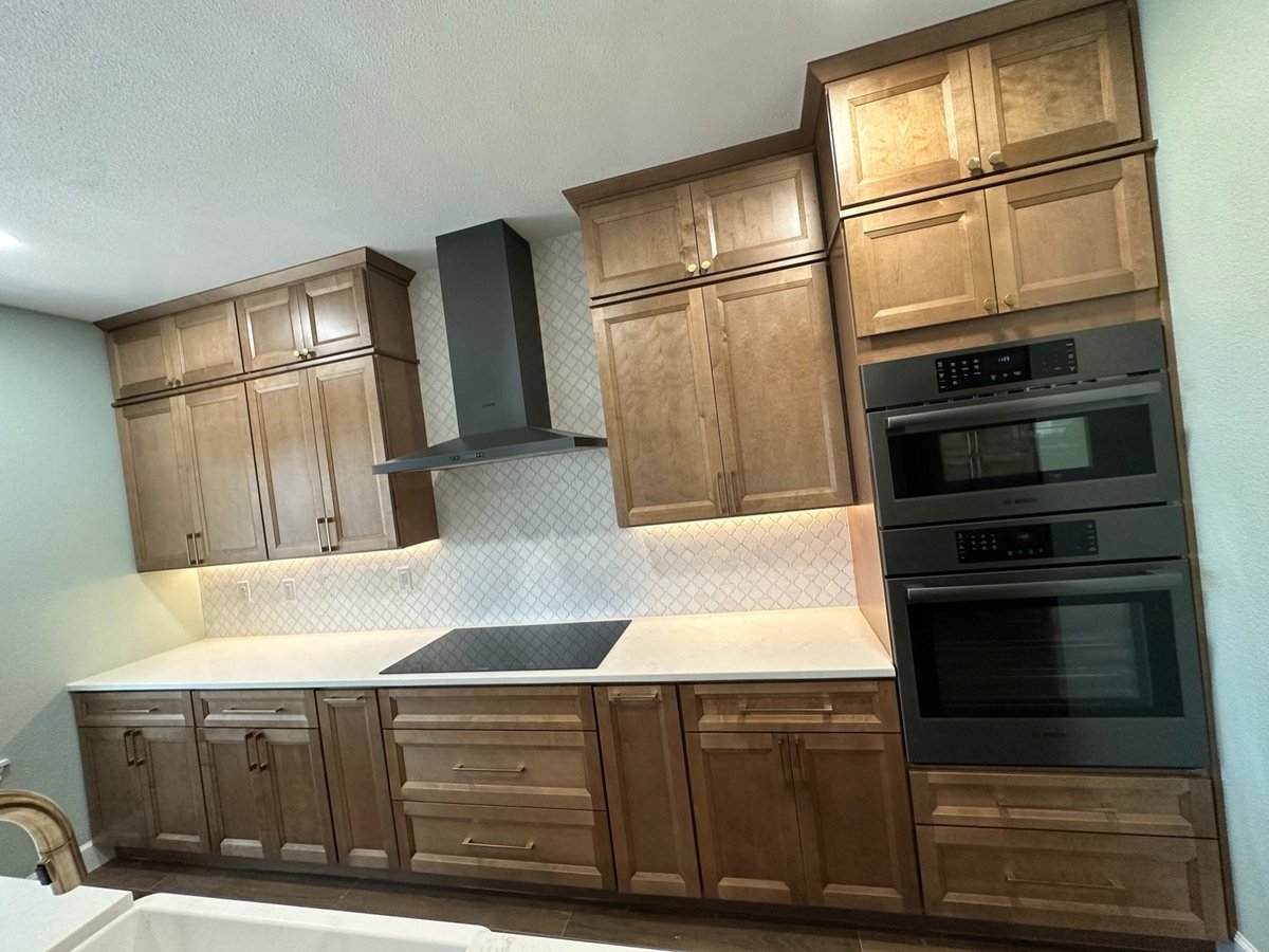 Stained cabinet run with cooktop, chimney hood and stacked black Bosch ovens