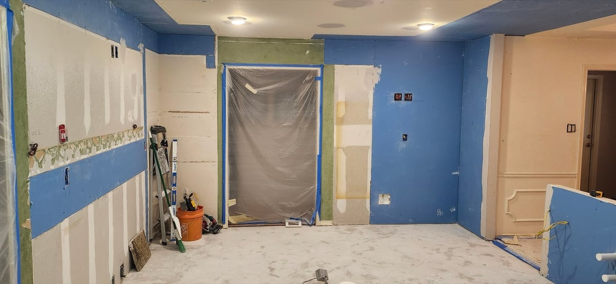 Gutted kitchen mid-remodel with exposed blue drywall and plastic-draped opening