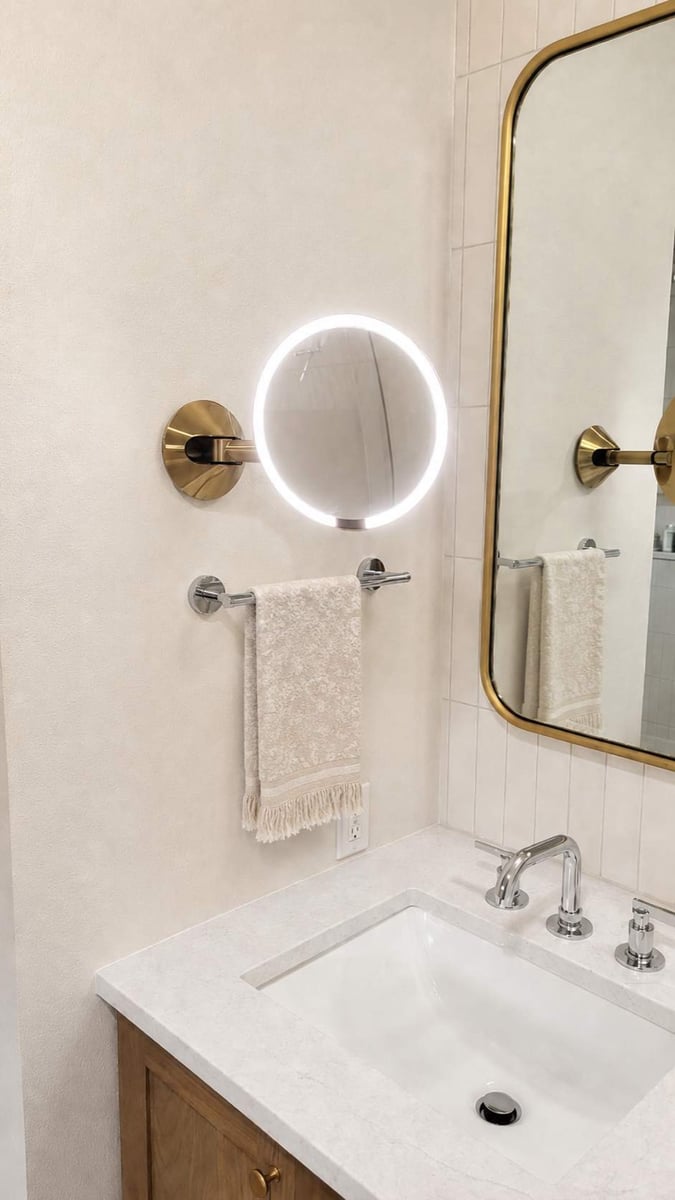 Lit round magnifying mirror and towel bar beside gold-framed mirror over white sink