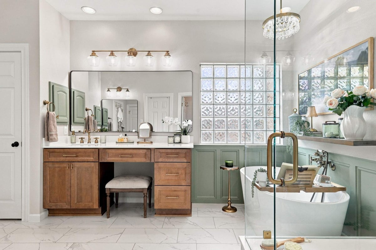 Rogers Ranch primary bathroom featuring a warm wood vanity with brass hardware, freestanding soaking tub, sage-green wainscoting, and glass-block window.