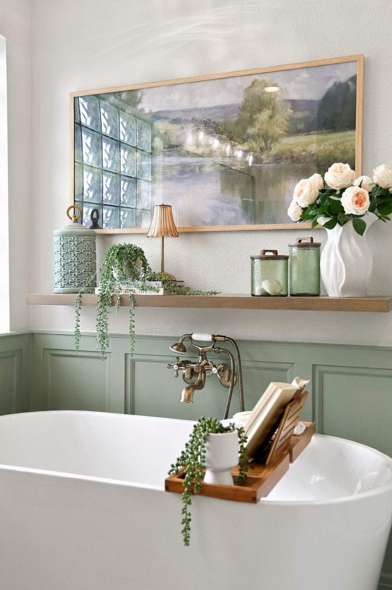 Freestanding white soaking tub against sage-green wainscoting in a Rogers Ranch bathroom, with brass fixtures, floating wood shelf, and decorative artwork.