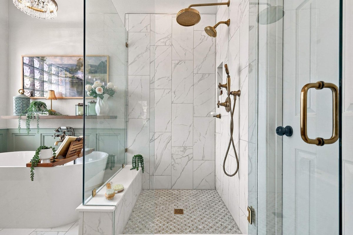Walk-in shower in a Rogers Ranch primary bathroom featuring marble-look wall tile, frameless glass enclosure, brass shower fixtures, and a decorative mosaic tile floor.