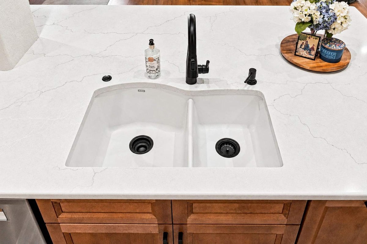 Double-basin kitchen sink with matte black faucet, white quartz-look countertop, and warm wood island cabinetry in a Cibolo Canyons kitchen remodel.