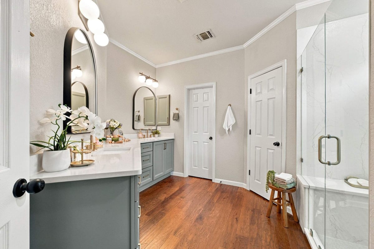 San Antonio bathroom remodel featuring sage-green double vanities, white countertops, arched mirrors, warm wood flooring, and a frameless glass walk-in shower.