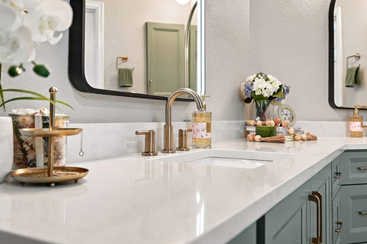 Close-up of a sage-green bathroom vanity with white countertops, brushed brass faucet, and black-framed mirrors in a San Antonio bathroom remodel.