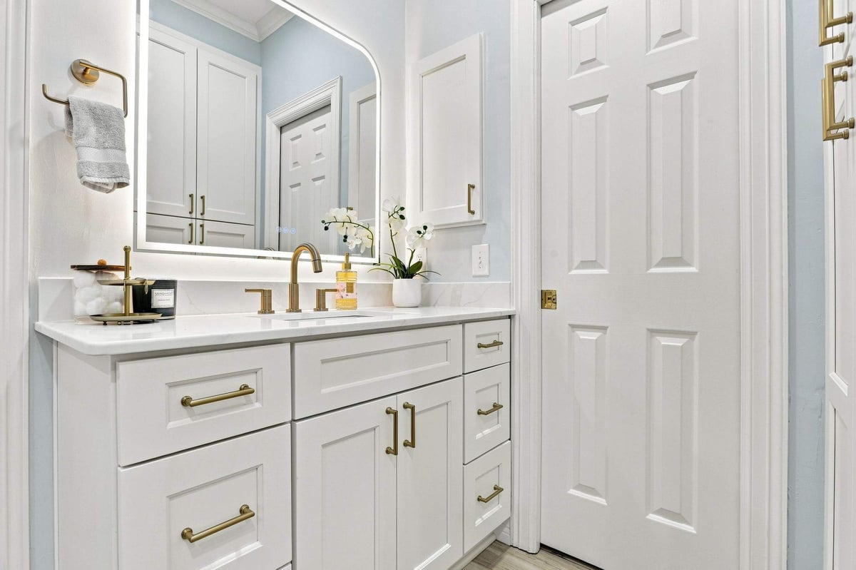 White bathroom vanity with quartz countertop, brushed-gold fixtures, and illuminated mirror.