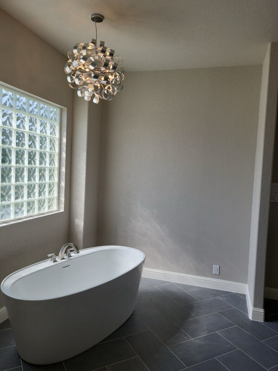 Freestanding white soaking tub below sculptural chandelier and glass block window