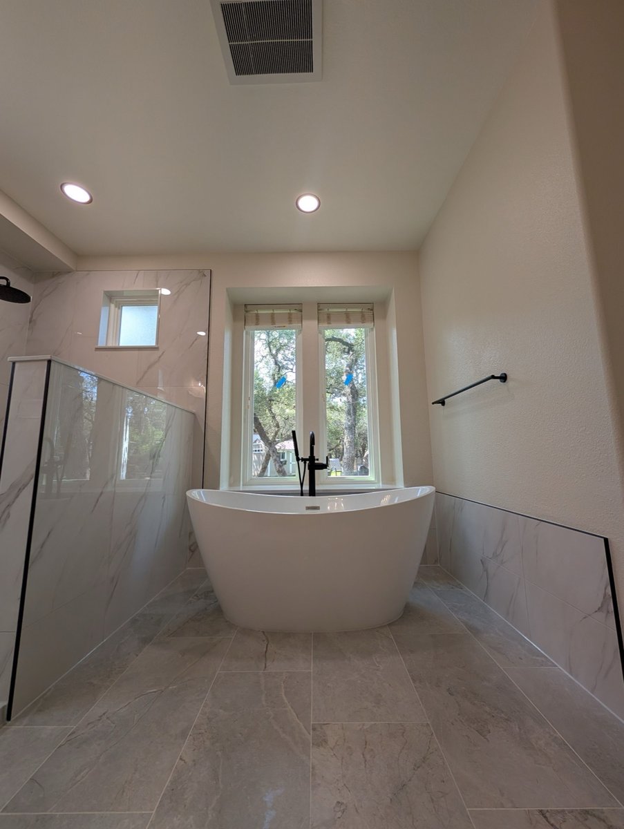 White freestanding tub under window between glass shower and marble wainscot