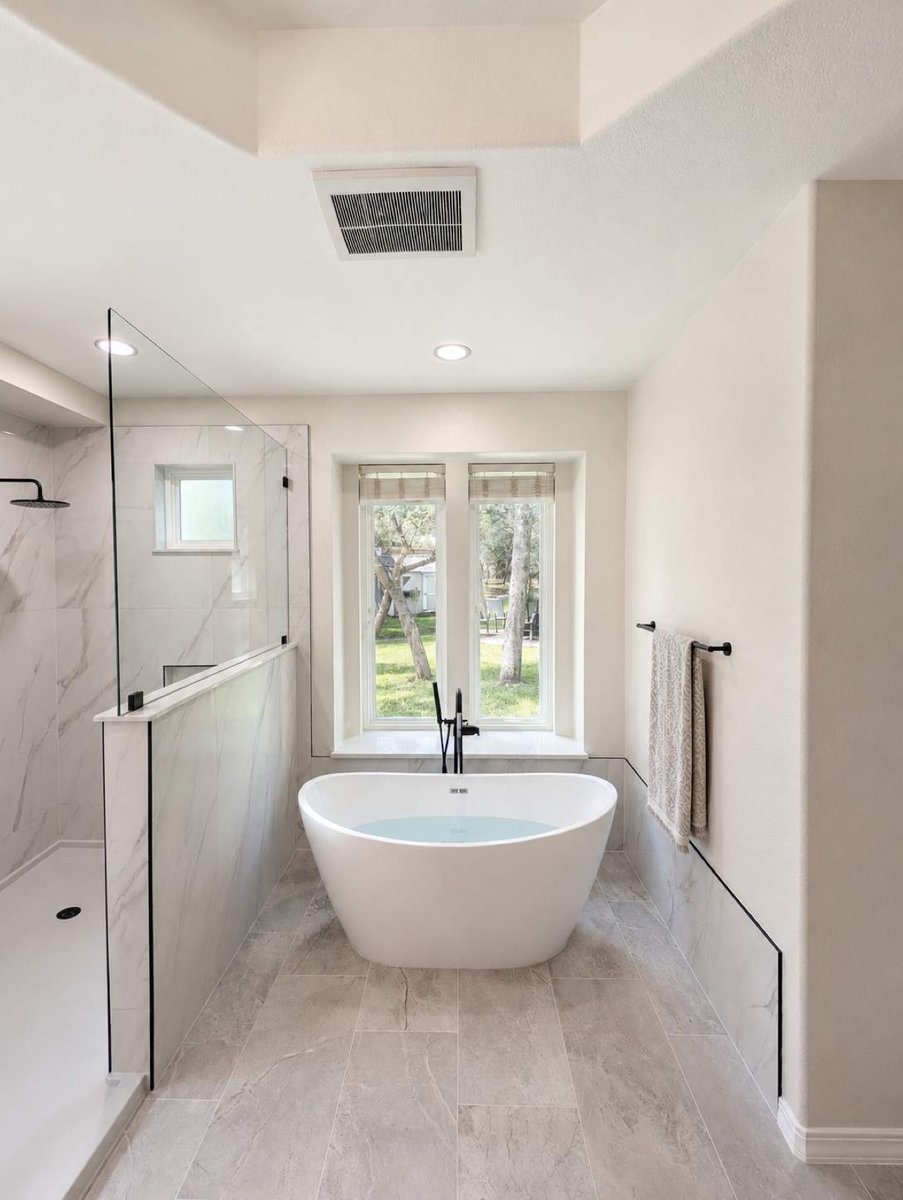 White soaking tub beneath windows beside marble half-wall glass shower