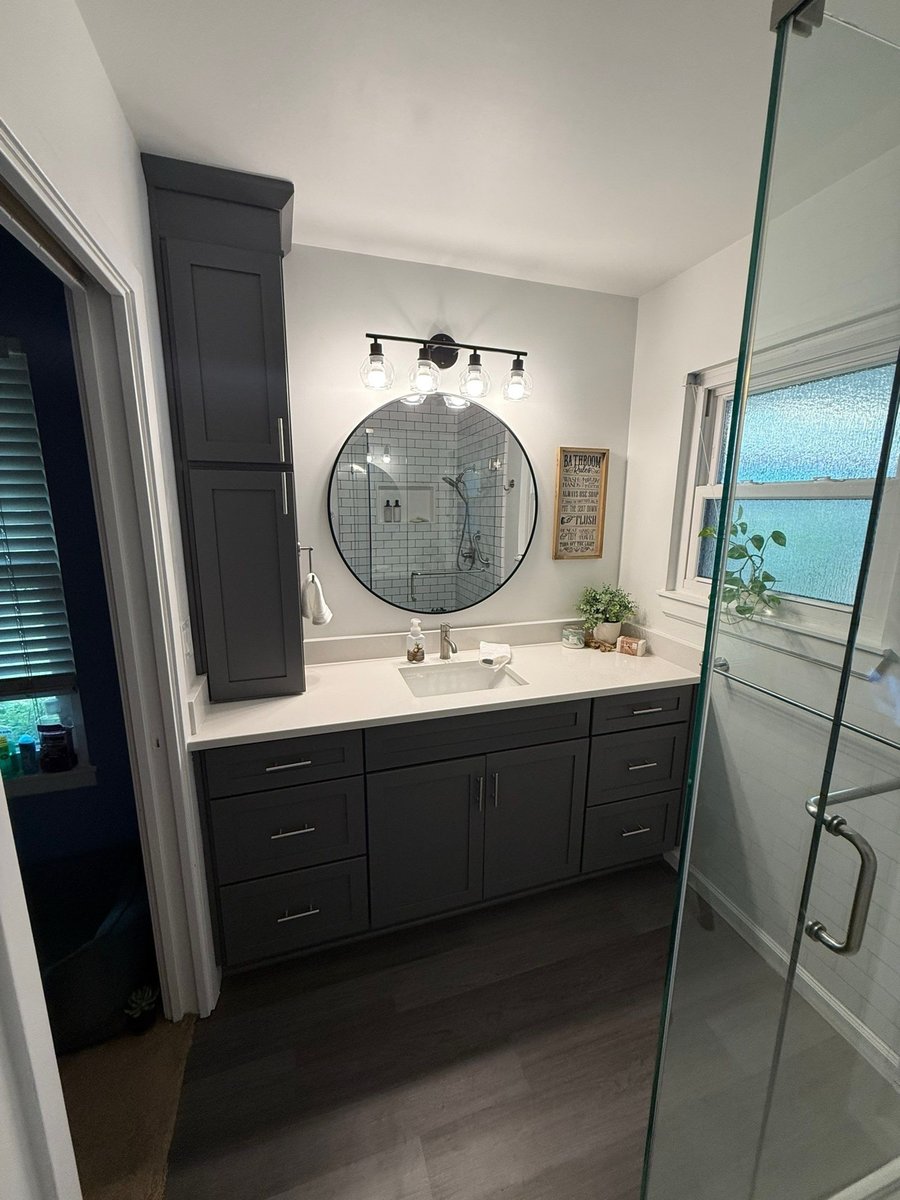 Gray vanity and round mirror with vanity bar beside glass shower