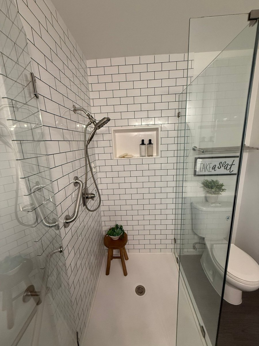 Subway-tiled corner shower with niche, wood stool and TAKE A SEAT sign