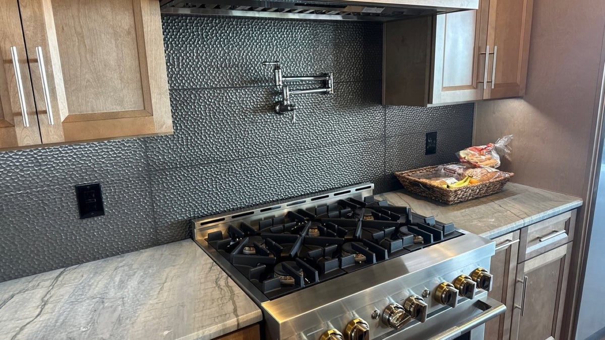 Stainless gas range below pot filler against a textured charcoal tile backsplash