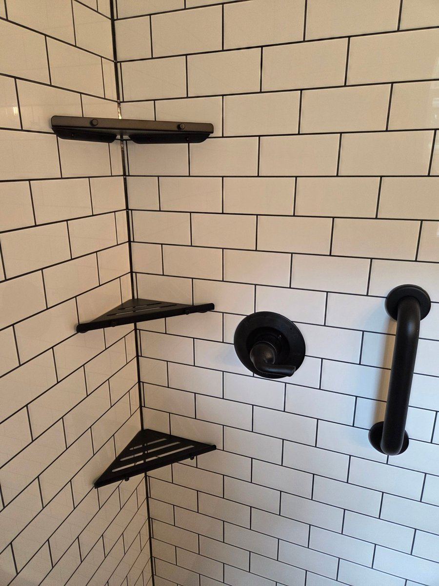 White subway tile shower corner with black shelves, grab bar, and water control