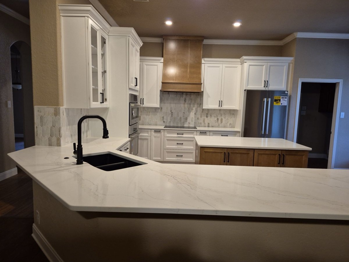 Wide marble peninsula counter with black sink facing white cabinets and wood hood