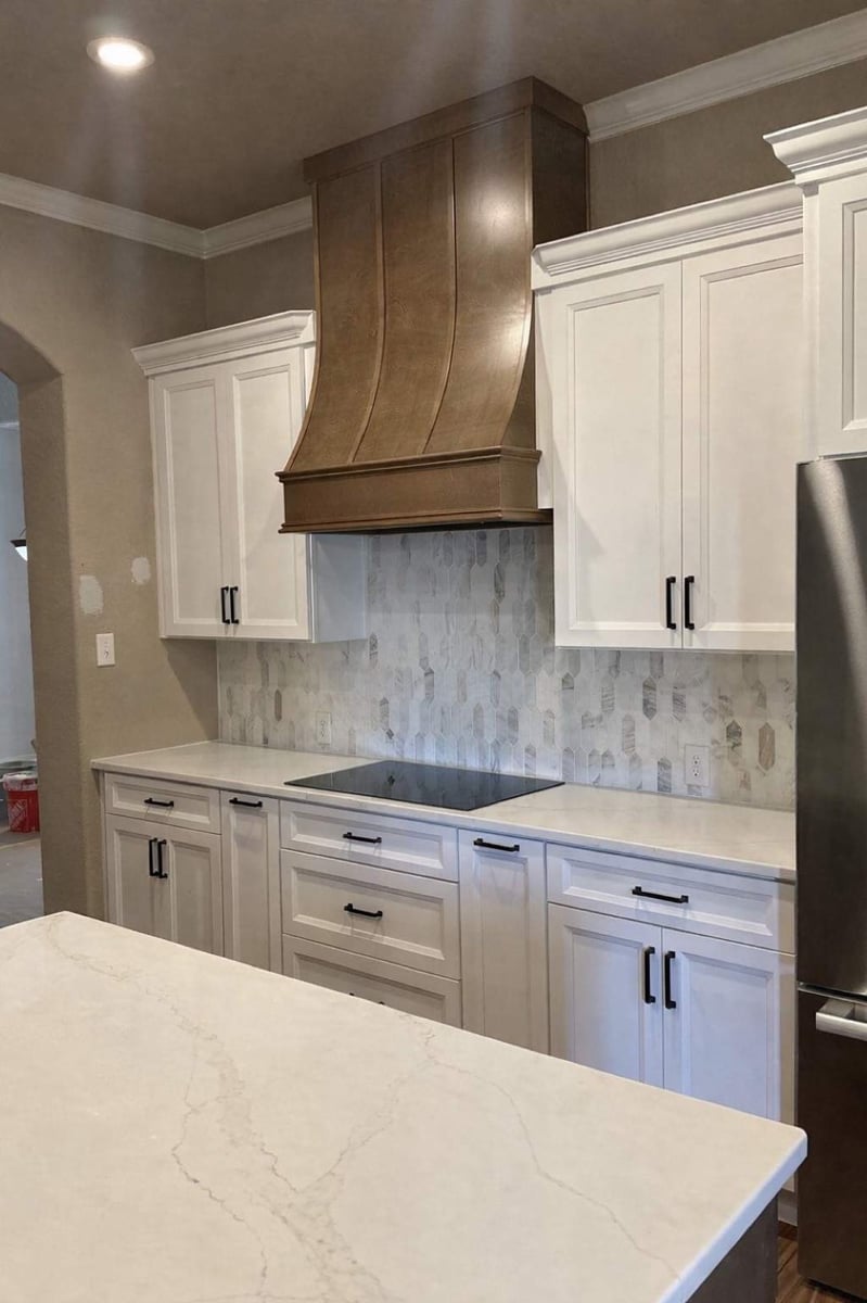 Wood range hood over cooktop with marble hexagon backsplash and white cabinets
