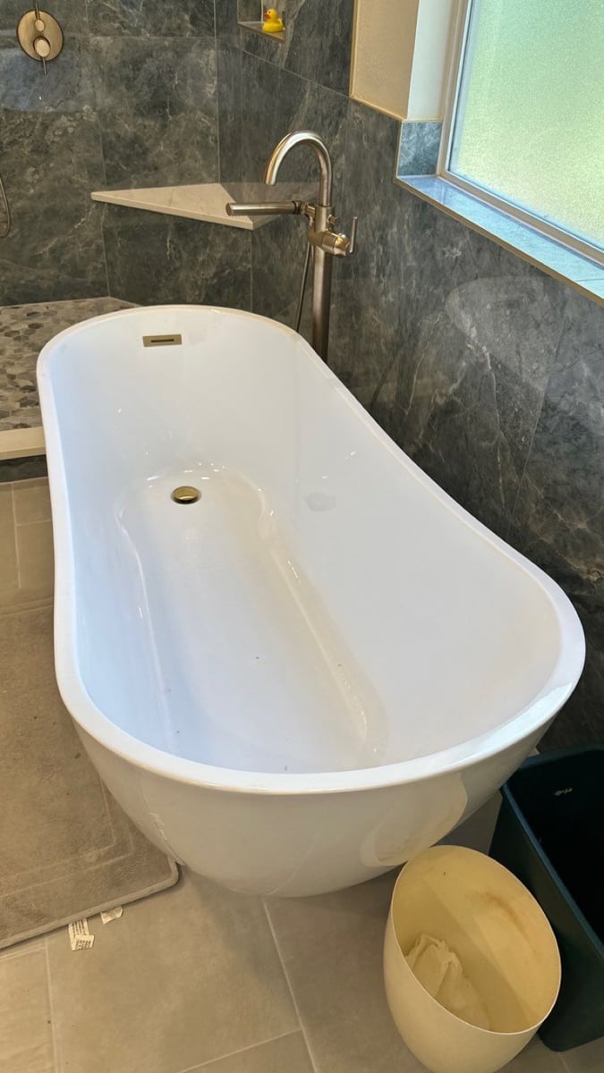 Freestanding white tub with brushed gold floor faucet against dark gray marble tile