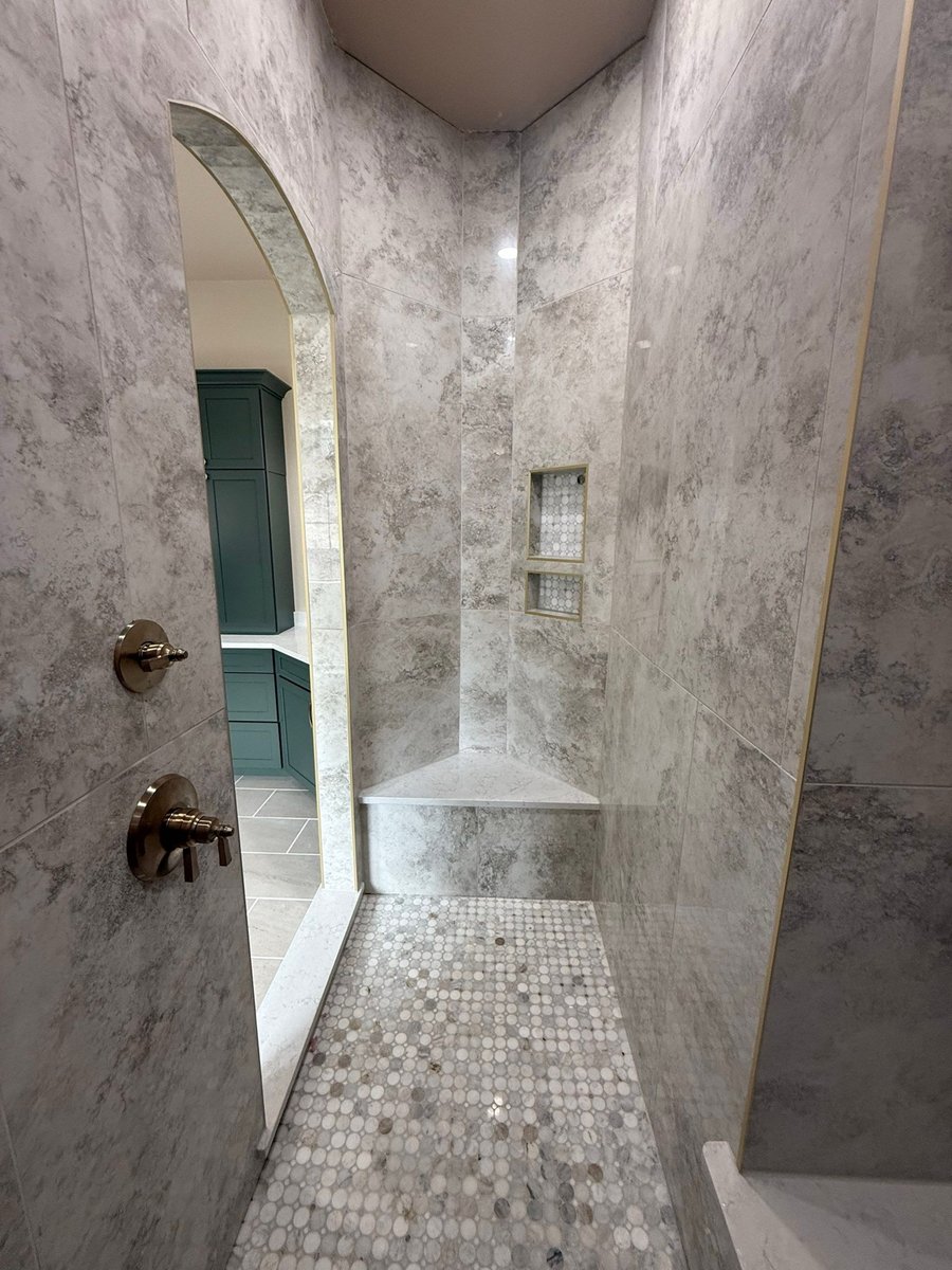 Arched shower entry with gray marble walls and corner bench, green cabinets beyond