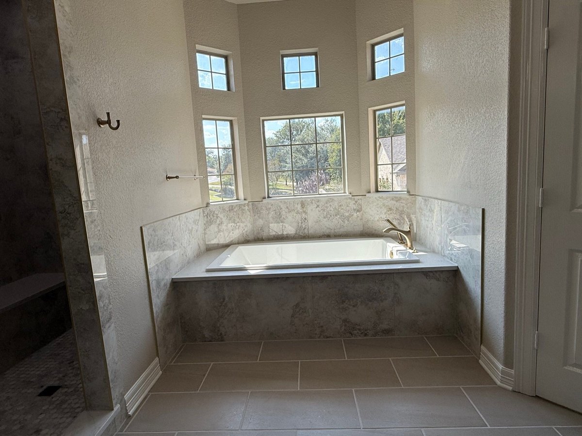 Drop-in soaking tub beneath three windows with marble surround and gray floor tile