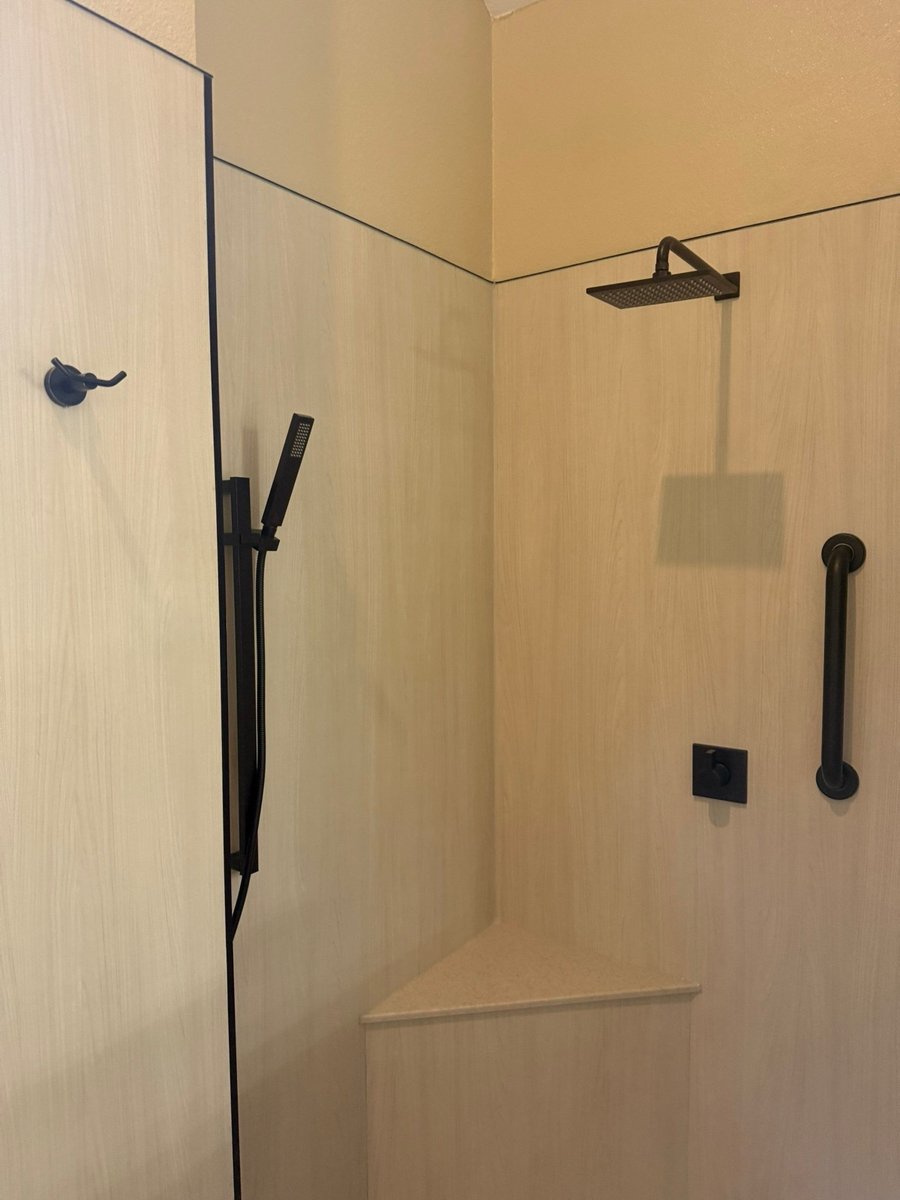 Curbless walk-in shower with light wood-grain panels, black rainfall head and corner seat