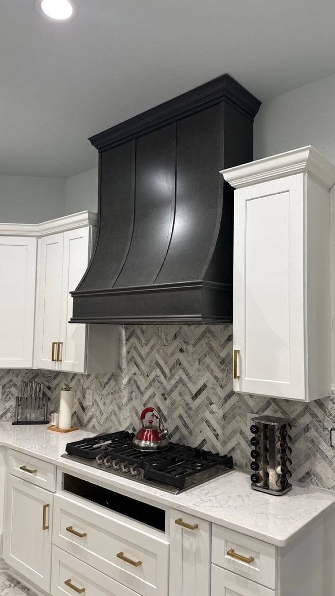 Black range hood over gas cooktop with gray herringbone marble backsplash and white shaker cabinets
