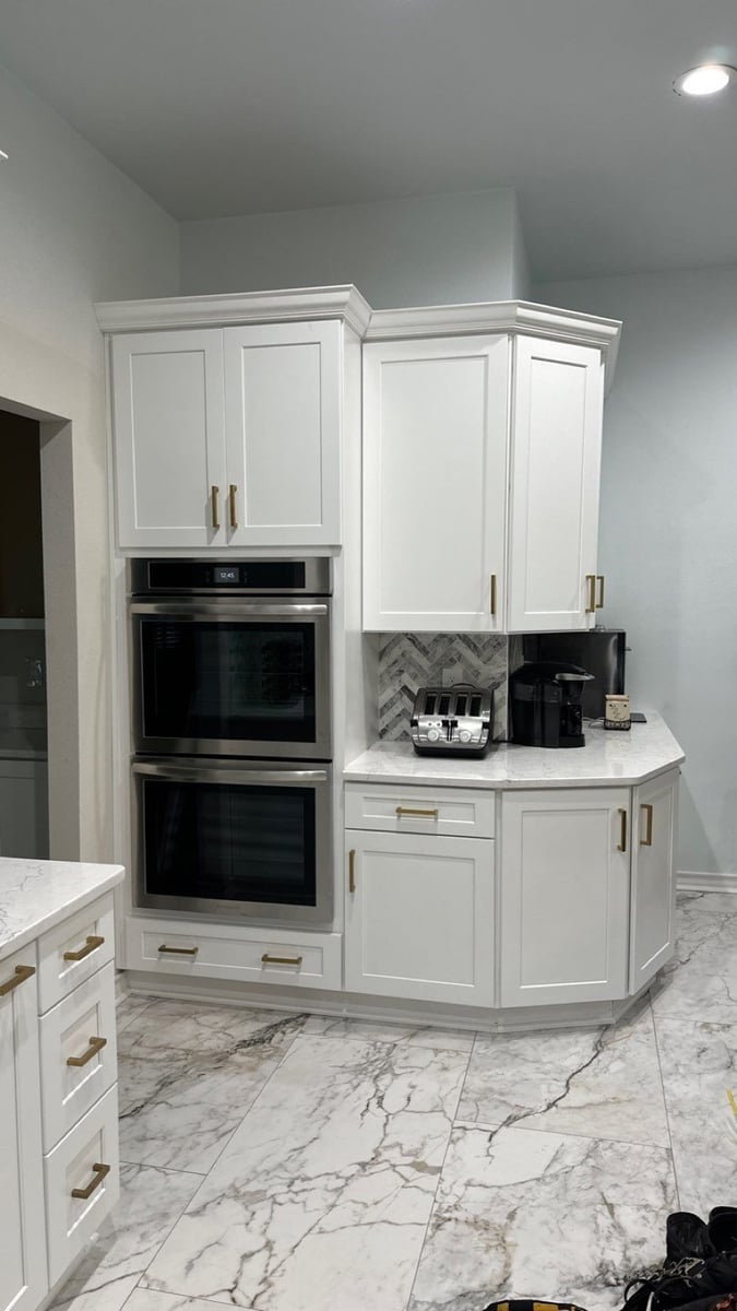 Corner of white kitchen with stainless double wall ovens and marble-look tile floor