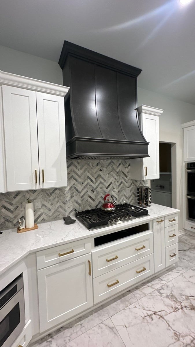 Gas cooktop below black range hood, gray herringbone backsplash, white cabinets, and marble countertops