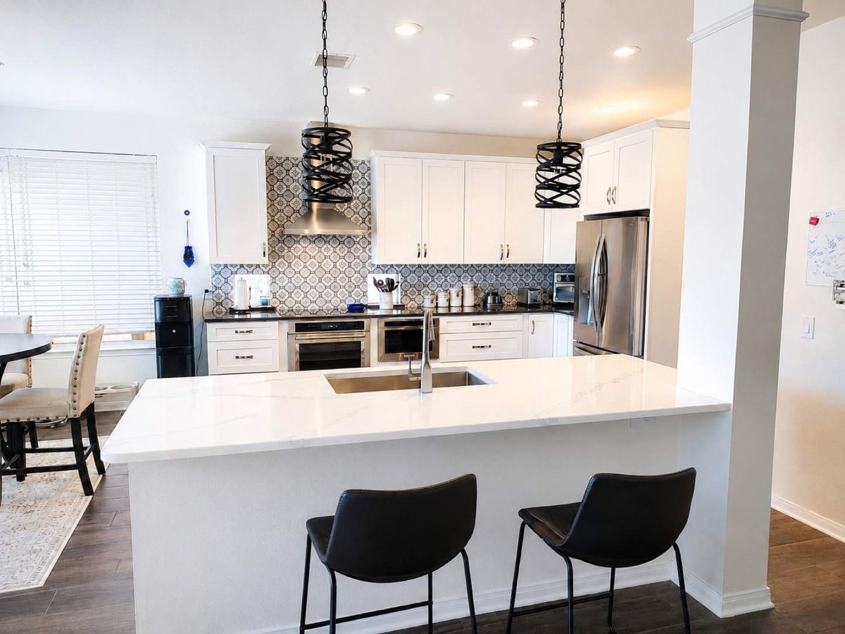 White kitchen with quartz peninsula, black barstools, blue patterned tile, and iron pendants