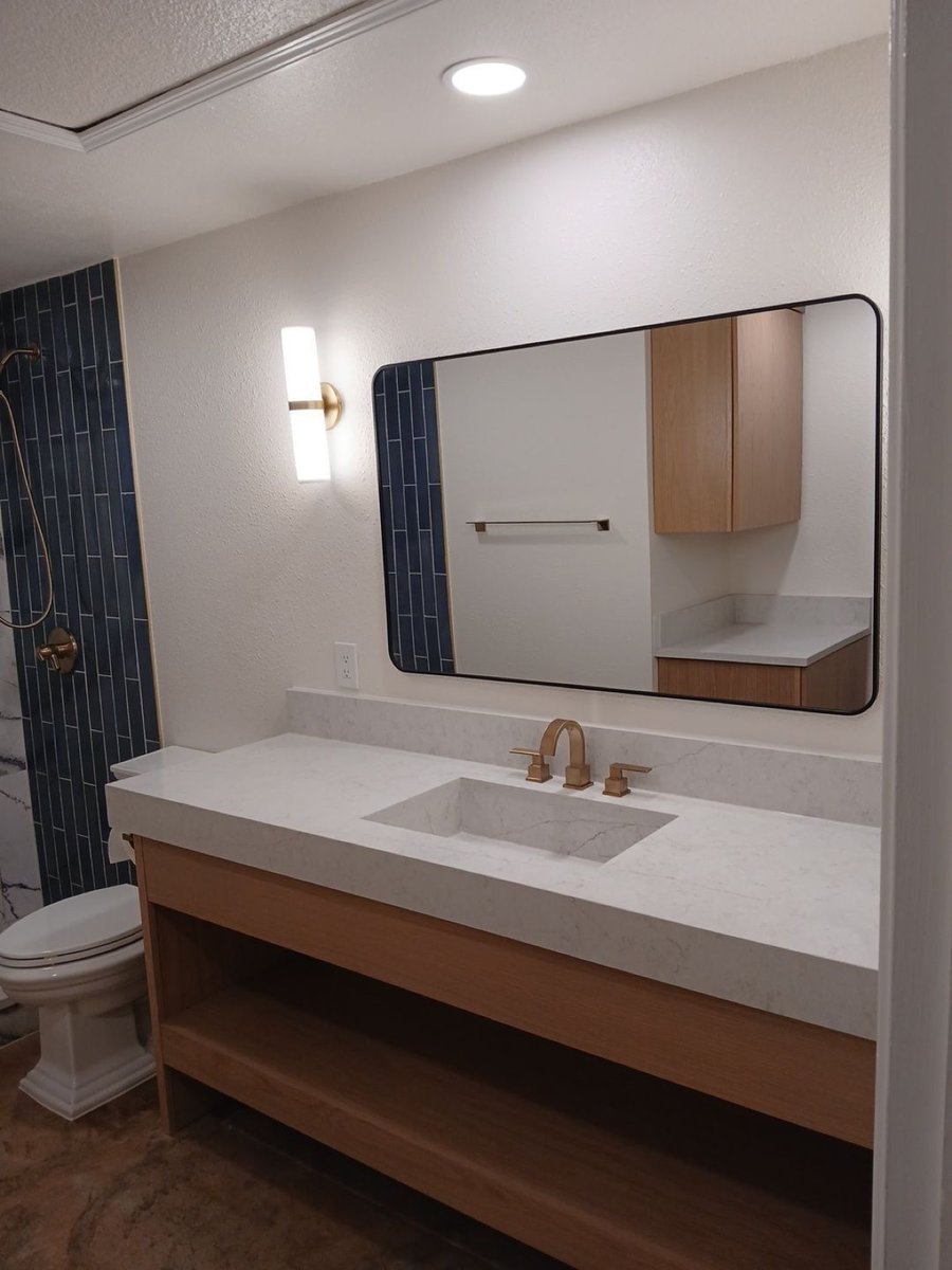Floating wood vanity with white quartz top, blue tile shower wall and brass faucet