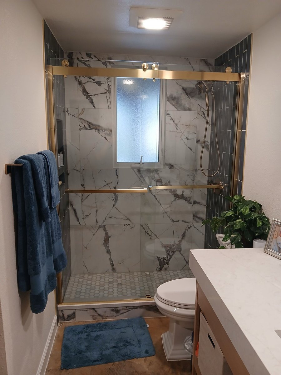 Glass sliding door shower with marble walls and gold frame beside white toilet