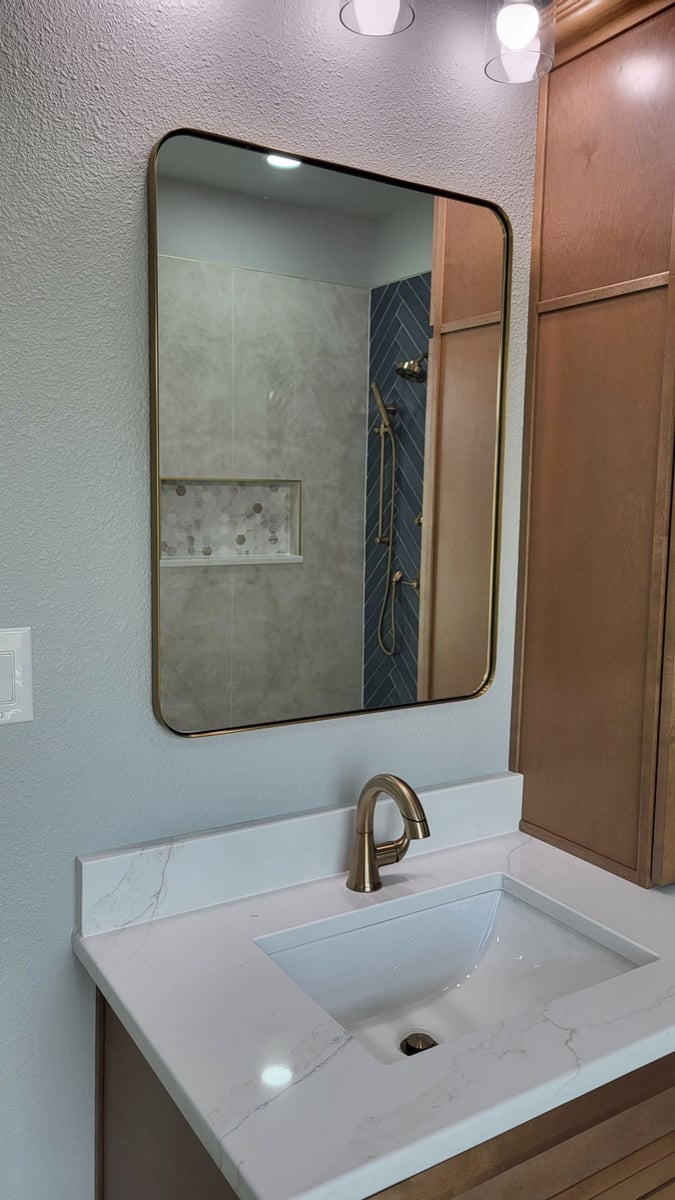 Wood vanity with white quartz top and gold-framed mirror reflecting blue tile shower