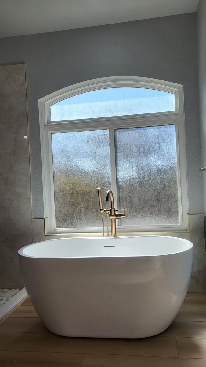 White freestanding tub below arched frosted window with brushed gold floor faucet
